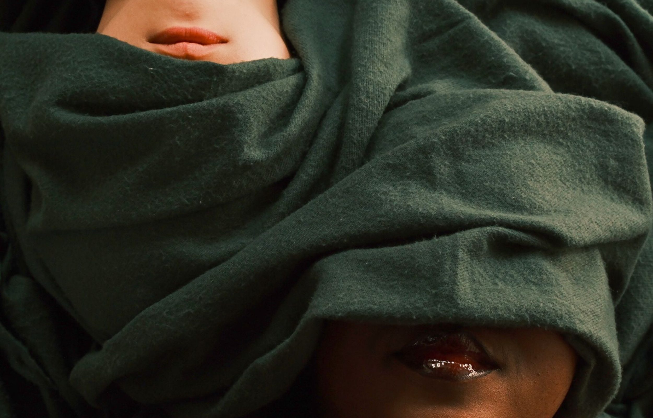 Two faces partially covered by a green cloth, with one face being white and the other black. Only their lips are visible, and the cloth obscures their eyes